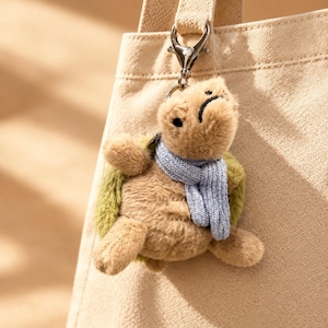 Pode incluir: Um porta-chaves de tartaruga de peluche com um corpo macio em tom castanho, uma carapaça verde texturizada e um pequeno cachecol de malha azul claro à volta do pescoço. A tartaruga está presa a um mosquetão de metal prateado num saco de lona bege.