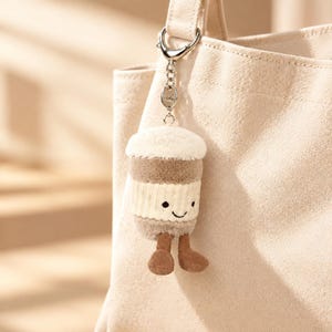 Pode incluir: Um porta-chaves de peluche em forma de copo de café com um topo creme, uma secção central castanha e uma faixa branca canelada com um rosto sorridente bordado. O porta-chaves tem duas pernas de veludo cotelê castanhas e um fecho prateado.