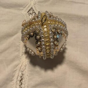 以下が含まれることがあります： 白いサテンのベースに、金色のスパンコールと小さな白いフェイクパールの縦帯が装飾された球形のオーナメント。上部には吊り下げ用の金色の金属製ループが付いており、精巧なビーズ細工が施された質感のあるデザインが特徴です。