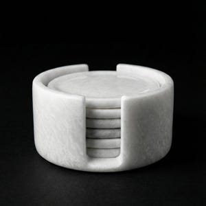 May include: A set of circular white marble coasters stacked neatly inside a matching cylindrical marble holder with a vertical cutout opening on the front side, showcasing the smooth, polished stone texture and consistent light grey veining.