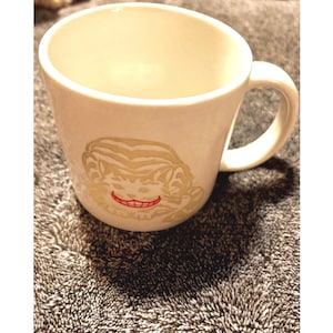 May include: A white ceramic mug featuring a light beige graphic of a cat face with striped fur patterns. The cat has a prominent, wide, red-coloured grin showing teeth. The mug has a smooth, rounded body and a single handle on the right side.