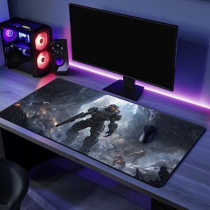 May include: A rectangular desk mat featuring a dark, detailed illustration of a futuristic armored soldier holding a rifle in a snowy, mountainous landscape. The mat is positioned on a desk surface beneath a computer monitor and a black mouse.