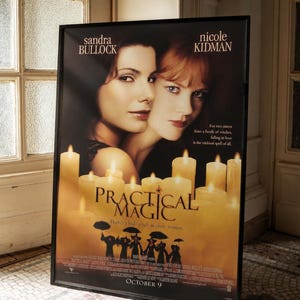 May include: A framed movie poster for Practical Magic featuring Sandra Bullock and Nicole Kidman. The poster displays the film title, cast names, and the tagline, There's a little witch in every woman, set against a background of glowing candles.