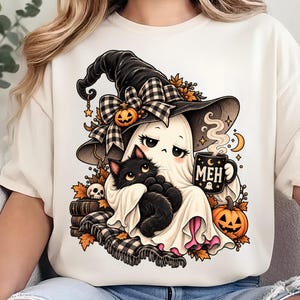Könnte beinhalten: Ein cremefarbenes T-Shirt mit einer Grafik eines Geistes, der einen schwarzen Hexenhut mit karierter Schleife trägt und eine schwarze Tasse mit der Aufschrift MEH und einem Halbmond hält. Eine schwarze Katze sitzt auf dem Schoß des Geistes, umgeben von Kürbissen.