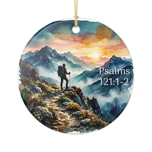 May include: A round ceramic ornament featuring a watercolor illustration of a hiker standing on a mountain trail at sunset. The scene includes blue peaks, misty valleys, and orange clouds. The text Psalms 121:1-2 is printed in white on the right.