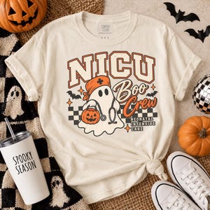 May include: A cream-coloured short-sleeve t-shirt featuring a graphic of a ghost wearing a medical scrub cap and stethoscope, holding a jack-o'-lantern. The text reads NICU Boo Crew Neonatal Intensive Care with a black and white checkered pattern.