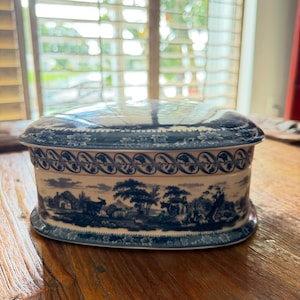 May include: An oval ceramic box with a white base and blue transferware pattern. The lid and sides feature detailed pastoral scenes of trees, livestock, and figures, accented by a decorative floral border along the top and bottom edges.