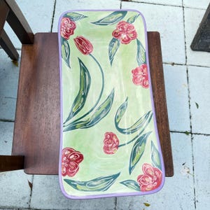 May include: A rectangular ceramic serving platter with rounded corners and a light purple rim. The surface features a light green background decorated with hand-painted red flowers and dark green leaves with white highlights.