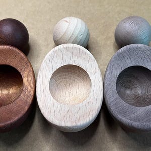 May include: Three wooden objects, each consisting of a smooth sphere resting atop an oval base with a central circular indentation. The set features one dark brown, one light beige, and one grey-toned wood piece with visible grain.