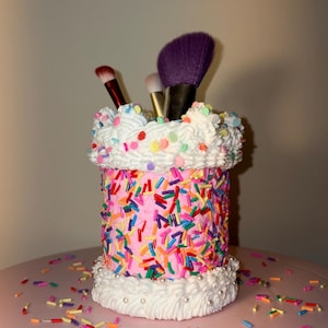 May include: A cylindrical makeup brush holder designed to resemble a pink frosted cake with white whipped cream borders at the top and bottom. The exterior is covered in colorful, multi-shaped sprinkles, and it holds three makeup brushes.