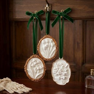 May include: Three oval-shaped hanging ornaments featuring white relief floral carvings of tulips, lily of the valley, and lilies. Each piece is encased in a decorative gold-toned frame and suspended by a dark green velvet ribbon bow.