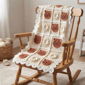 May include: A cream-colored crochet blanket featuring a grid of alternating squares. Some squares display a rust-colored cat face with embroidered features, while others contain a textured, circular floral motif. The blanket has a scalloped edge.