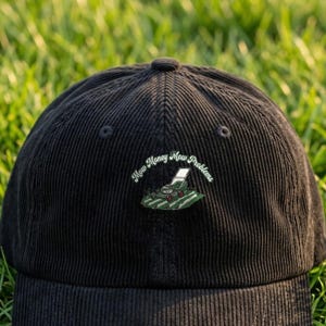 May include: A black corduroy baseball cap featuring an embroidered graphic of a green lawnmower on grass with the text "Mow Money Mow Problems" arched above the illustration in white lettering.
