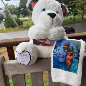 May include: A white plush teddy bear with dark-tipped ears and a black nose. The bear holds a white fabric panel featuring a printed photograph of two people in a pool. A heart-shaped patch on the foot reads: August 22, 2026, Nicole & T.J., Engaged.