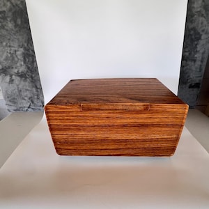 May include: A rectangular wooden box with a rich, dark brown wood grain pattern. The box features a smooth, flat lid and a horizontal wooden handle centered on the front panel, showcasing a natural, polished finish throughout the exterior.