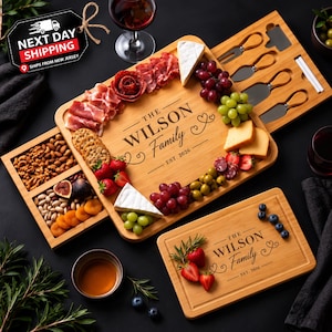 May include: A bamboo charcuterie board set featuring a slide-out drawer with four cheese knives, engraved with "THE WILSON Family EST. 2026". The board is arranged with assorted meats, cheeses, grapes, strawberries, nuts, and dried fruit.