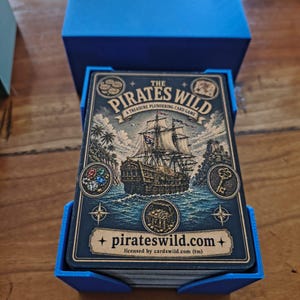 May include: A deck of The Pirates Wild card game in a blue plastic holder with a matching blue lid. The card box features a detailed illustration of a pirate ship at sea, a treasure chest, a key, and jewels, with the text The Pirates Wild.