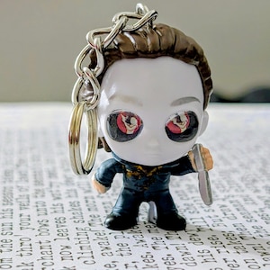 May include: A keychain featuring a small figurine of a character with brown hair, a white mask, and a dark blue jumpsuit. The character has large black eyes with red circular accents and holds a small silver-colored knife in one hand.
