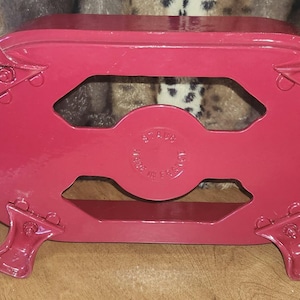 May include: A glossy red enamelled cast iron trivet with an oval frame, four curved legs, and side handles. The centre features a circular embossed logo that reads Staub Made in France, providing a stable base for hot cookware in the kitchen.