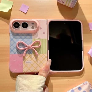 May include: A foldable smartphone case in pastel pink, blue, and green with a patchwork pattern, a raised camera housing, and a three-dimensional pink bow accent. The case features the word LUCKY printed in small letters on the lower section.