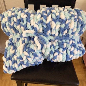 May include: A chunky knit blanket featuring a variegated pattern of royal blue, light blue, and white yarn. The thick, soft-textured throw is folded and secured in the center with a matching light blue yarn tie, creating a bundled appearance.