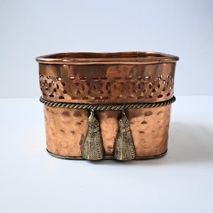 May include: A rectangular copper container with rounded corners, featuring a hammered lower section and a decorative pierced upper band. A twisted metal rope trim encircles the middle, adorned with two hanging metal tassels.