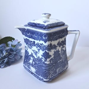 May include: A square-shaped ceramic teapot featuring a blue and white willow pattern. The design depicts pagodas, bridges, and trees. It includes a matching lid with a small knob handle and a side handle. The teapot has a white background.