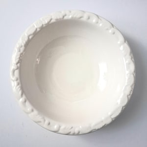 May include: A round, off-white ceramic bowl featuring a smooth, glossy interior and a raised, textured relief pattern along the outer rim. The bowl has a deep, circular basin and a wide, decorative edge that encircles the entire top circumference.