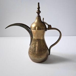 May include: A brass coffee pot featuring a long, curved spout, a hinged lid with a pointed finial, and a slender handle. The body of the pot is decorated with an etched floral pattern, showcasing a metallic gold finish throughout the piece.