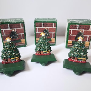 May include: Three green cast iron Christmas tree stocking hangers, each topped with a small angel figure. The trees are decorated with white stars, red ornaments, and a toy train at the base. Each hanger is accompanied by its original green box.