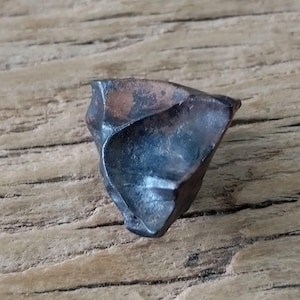 Puede incluir: Un pequeño fragmento de piedra de forma irregular, oscuro, con un brillo metálico y parches de color cobre visibles. La superficie muestra bordes afilados y angulares con una textura suave y reflectante, destacando un contraste marcado entre las áreas de color gris carbón y bronce en su forma compacta y multifacética.