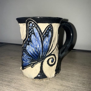 May include: A ceramic mug with a textured cream-colored surface featuring a prominent blue butterfly design. The mug has a black rim, a black handle, and black accents surrounding the butterfly, with carved details throughout the exterior.