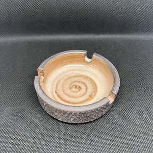 May include: A round ceramic ashtray featuring a textured dark gray exterior and a smooth, light beige interior basin. The center of the basin displays a subtle, hand-painted brown spiral pattern, and the rim includes three evenly spaced notches.