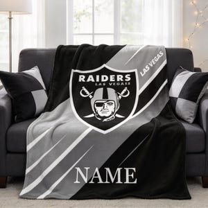 May include: A black and grey fleece blanket featuring the Las Vegas Raiders logo with a shield, a pirate mascot, and crossed swords. The text reads Raiders Las Vegass, Las Vegas, and Name. The design includes diagonal stripes across the fabric.
