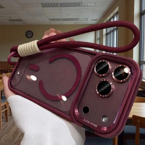 May include: A deep maroon smartphone case featuring a circular magnetic ring on the back, a triple camera cutout with metallic rose gold rims, and a matching maroon braided wrist strap attached with a cream-colored cylindrical connector.