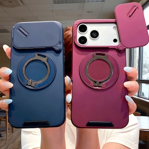 May include: Two protective smartphone cases, one in dark blue and one in deep red, each featuring a sliding camera lens cover, a circular ring stand on the back, and a black accent piece at the bottom edge of the case for added durability.