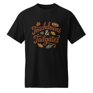 Puede incluir: Una camiseta negra de manga corta con el texto Touchdowns & Tailgates en una fuente cursiva marrón. El diseño incluye un gráfico de un balón de fútbol americano marrón y varias hojas de colores otoñales en tonos naranja, amarillo y beige.