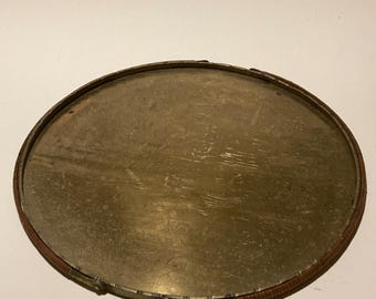 May include: An oval, antique brass tray with a woven brown border. The tray's surface has a worn, aged appearance, with subtle patterns and markings. The rim is a darker brown, adding contrast to the brass.