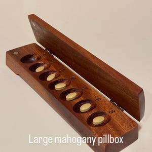 Puede incluir: Un pastillero grande de caoba abierto, con seis compartimentos ovalados, cada uno con una pastilla amarilla. La tapa está abierta, revelando el interior. El texto "Large mahogany pillbox" es visible.