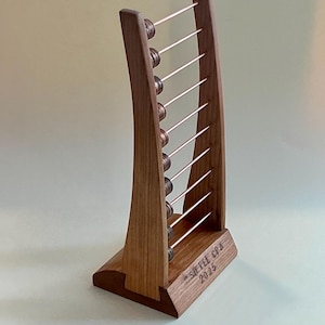 Puede incluir: Un ábaco de madera con un marco curvo y varillas de color cobre. El ábaco tiene diez filas de cuentas y una base con el texto "SIEPEL CPA 2025" grabado. La madera tiene un tono cálido y natural.