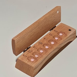May include: Wooden pill organizer with a hinged lid. The organizer is made of light-colored wood and features seven compartments with pink pill-shaped inserts. The days of the week are labeled on the front.