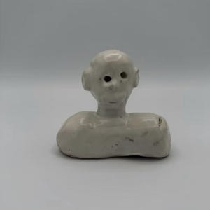 May include: A small, off-white ceramic figurine featuring a stylized head with prominent ears and circular eye cutouts, attached to a smooth, elongated, rectangular base. The surface has a glossy, glazed finish with subtle grey speckles.