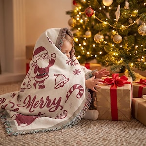 May include: A cream-colored woven throw blanket featuring a maroon pattern of Santa Claus, bells, candy canes, snowflakes, and the text Merry Christmas, with fringed edges, draped over a child reaching toward a gift box with a red ribbon.
