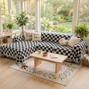 May include: A sectional sofa covered with a black and white checkered throw blanket featuring fringe edges. The sofa includes two cream-colored square pillows with the text LOVE HOME printed in black, and one cream-colored knot pillow.
