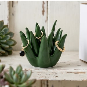 May include: A glossy dark green ceramic ring holder shaped like a succulent plant with multiple pointed leaves. Two gold-toned rings are placed on the leaves, one featuring a small black circular charm and the other with a textured band.