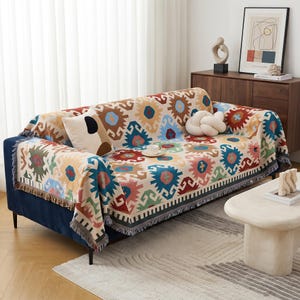 May include: A rectangular woven throw blanket featuring a repeating geometric pattern in shades of red, blue, green, and tan, with fringed edges, draped over a dark blue sofa. The blanket displays a series of stylized, multi-colored shapes.
