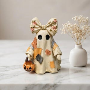 May include: A cream-coloured ghost figurine featuring a large bow on its head decorated with small pink roses. The ghost is wearing a patchwork-style robe with orange, grey, and tan fabric patches and is holding a small orange jack-o'-lantern bucket.