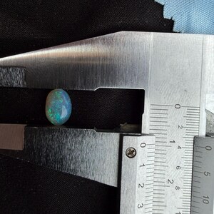 May include: An oval-shaped opal gemstone with iridescent flashes of green and blue color. The stone is positioned between the jaws of a metal caliper, which indicates a length of approximately 0.35 inches for the gemstone.