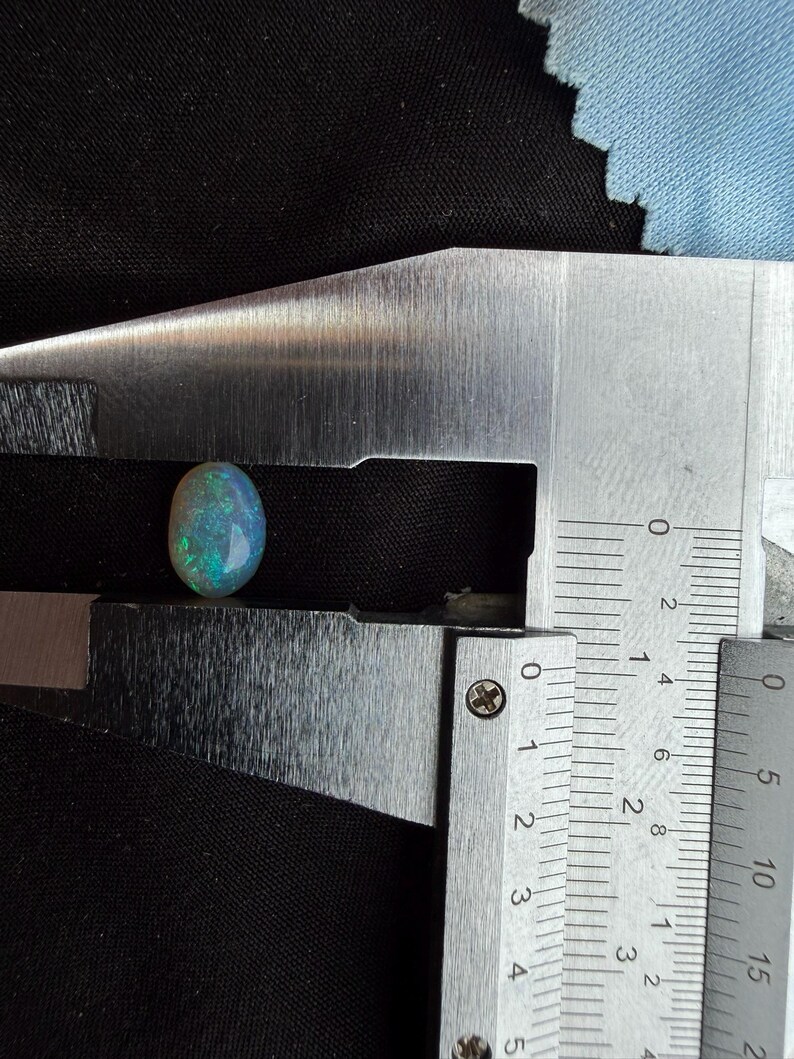 May include: An oval-shaped opal gemstone with iridescent flashes of green and blue color. The stone is positioned between the jaws of a metal caliper, which indicates a length of approximately 0.35 inches for the gemstone.