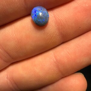 May include: A small, oval-shaped black opal gemstone featuring a dark base color with vibrant, iridescent flashes of bright blue and deep violet light reflecting across its polished, smooth, and rounded surface.